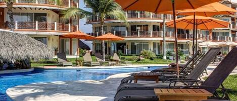 A resort pool area with lounge chairs and umbrellas, a thatched structure, and multi-story buildings with balconies.