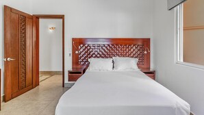 Habitación de hotel con cabecera de madera, cama con ropa de cama blanca, una mesita de noche y una puerta que da a otra habitación.