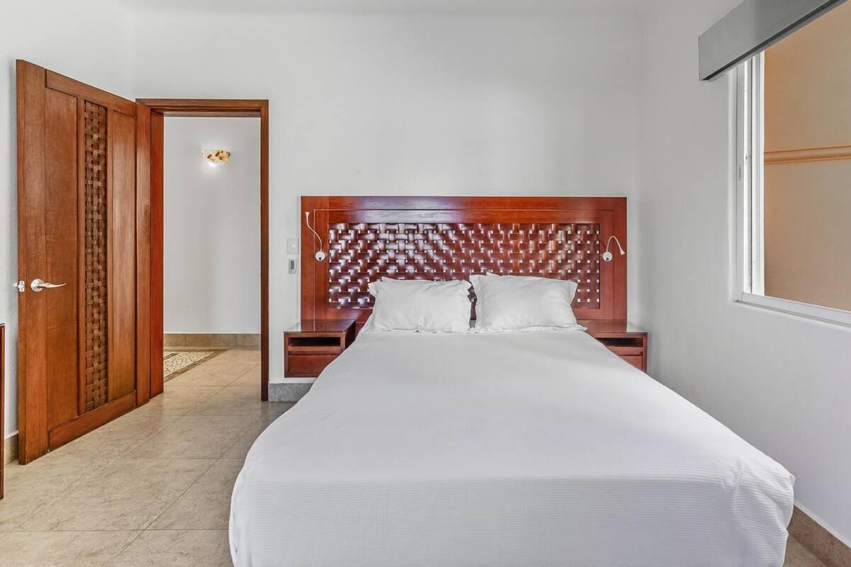 A hotel room with a wooden headboard, a bed with white bedding, a nightstand, and a door leading to another room.