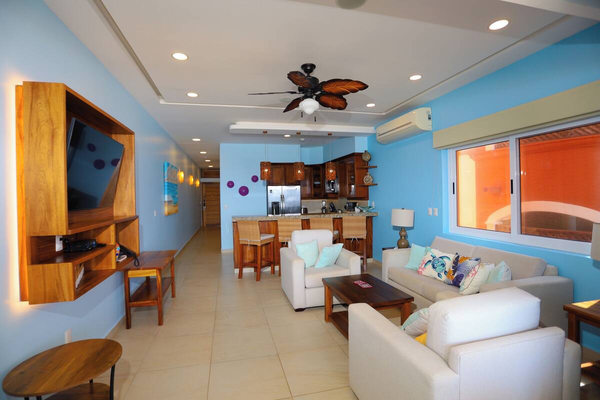 A modern living room with a kitchen in the background, a ceiling fan, and a TV mounted on the wall.