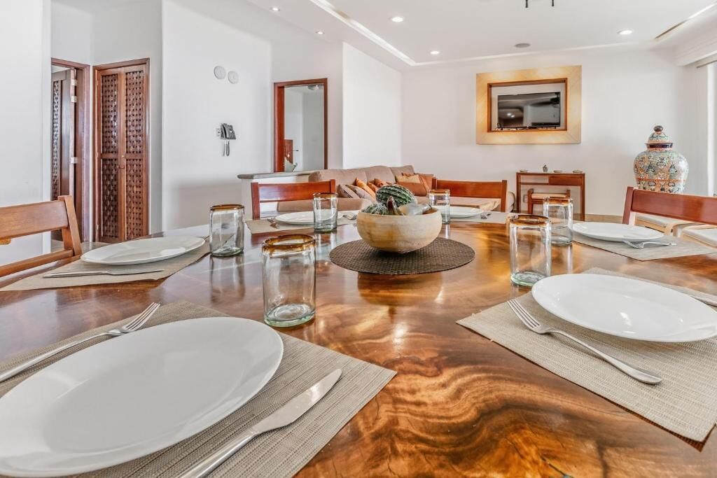 A dining room with a wooden table set for six, surrounded by chairs, with a large vase and a bowl on the table.