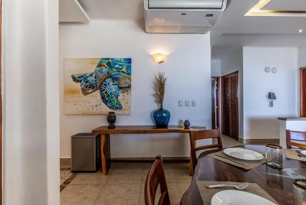 A dining area with a wooden table, chairs, a painting, a vase, and a small refrigerator.