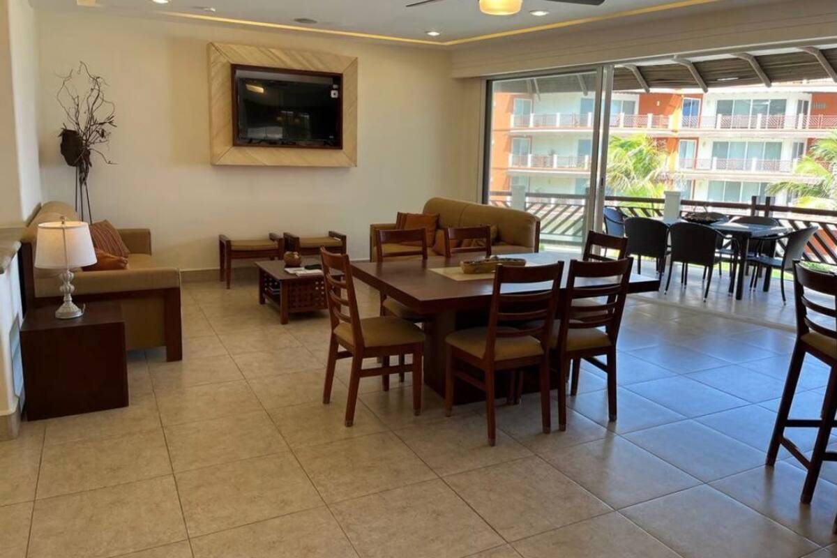 A spacious living area with a dining table, chairs, sofas, a TV mounted on the wall, and a balcony with a view of buildings and greenery.