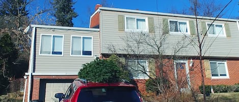 Un immeuble résidentiel de deux étages avec un garage, une voiture rouge garée devant et un sapin de Noël sur le toit.