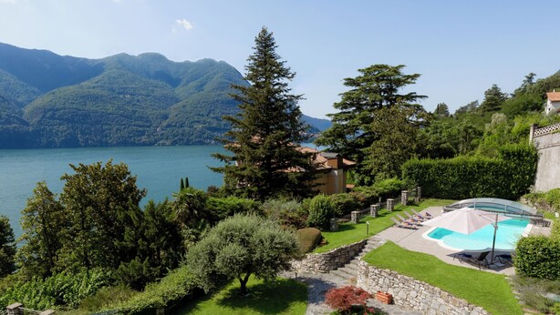 Un rigoglioso giardino con vialetto in pietra, una piscina e vista su un lago e montagne.