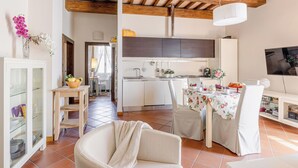 Una cocina moderna con armarios blancos, un comedor con una mesa dispuesta para dos personas y una sala de estar con sofá y televisor.