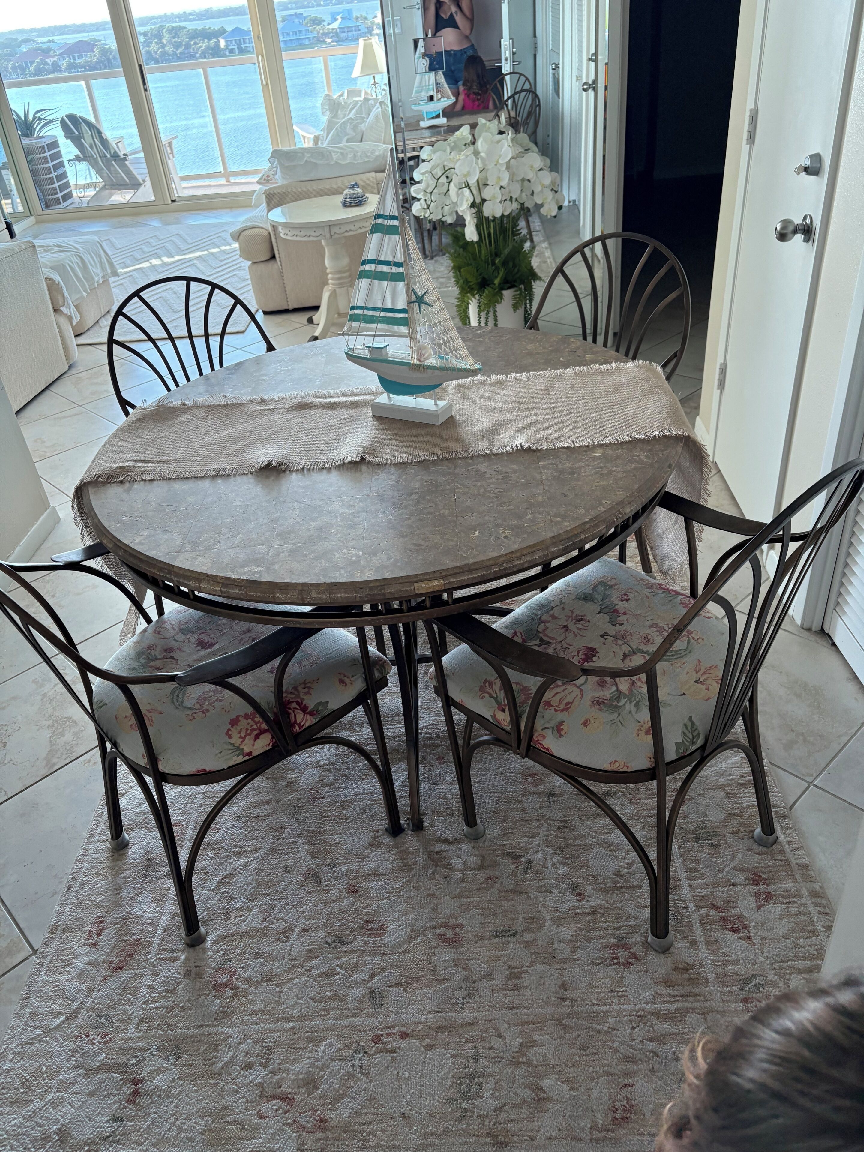 A round dining table with chairs, a small sailboat decoration, and a view of the sea through large windows.