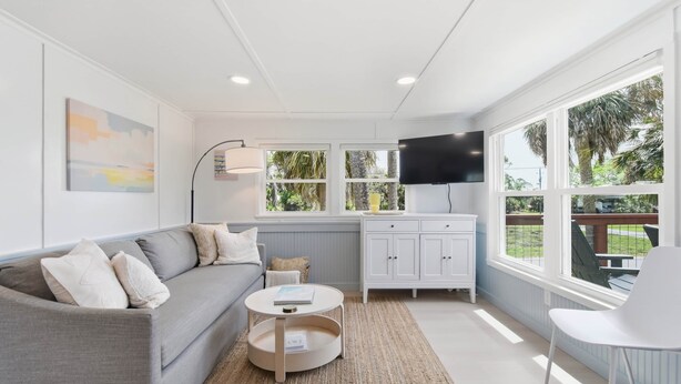 A modern living room with a grey sofa, a round coffee table, a white cabinet, a flat-screen TV, and large windows with a view of greenery. - Seabreeze Cottage - Home in Steinhatchee Florida (Steinhatchee)