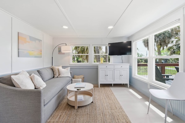A modern living room with a grey sofa, a round coffee table, a white cabinet, a flat-screen TV, and large windows with a view of greenery. - Seabreeze Cottage - Home in Steinhatchee Florida (Steinhatchee)