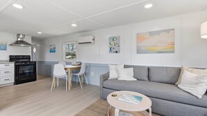 A modern living room with a grey sofa, a small round coffee table, a dining area with a white chair, and a kitchen with a black stove. - Seabreeze Cottage - Home in Steinhatchee Florida (Steinhatchee)