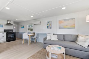 A modern living room with a grey sofa, a small round coffee table, a dining area with a white chair, and a kitchen with a black stove. - Seabreeze Cottage - Home in Steinhatchee Florida (Steinhatchee)