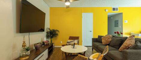 A living room with a ceiling fan, a flat-screen TV, a round coffee table, a sofa, and a wooden chair.