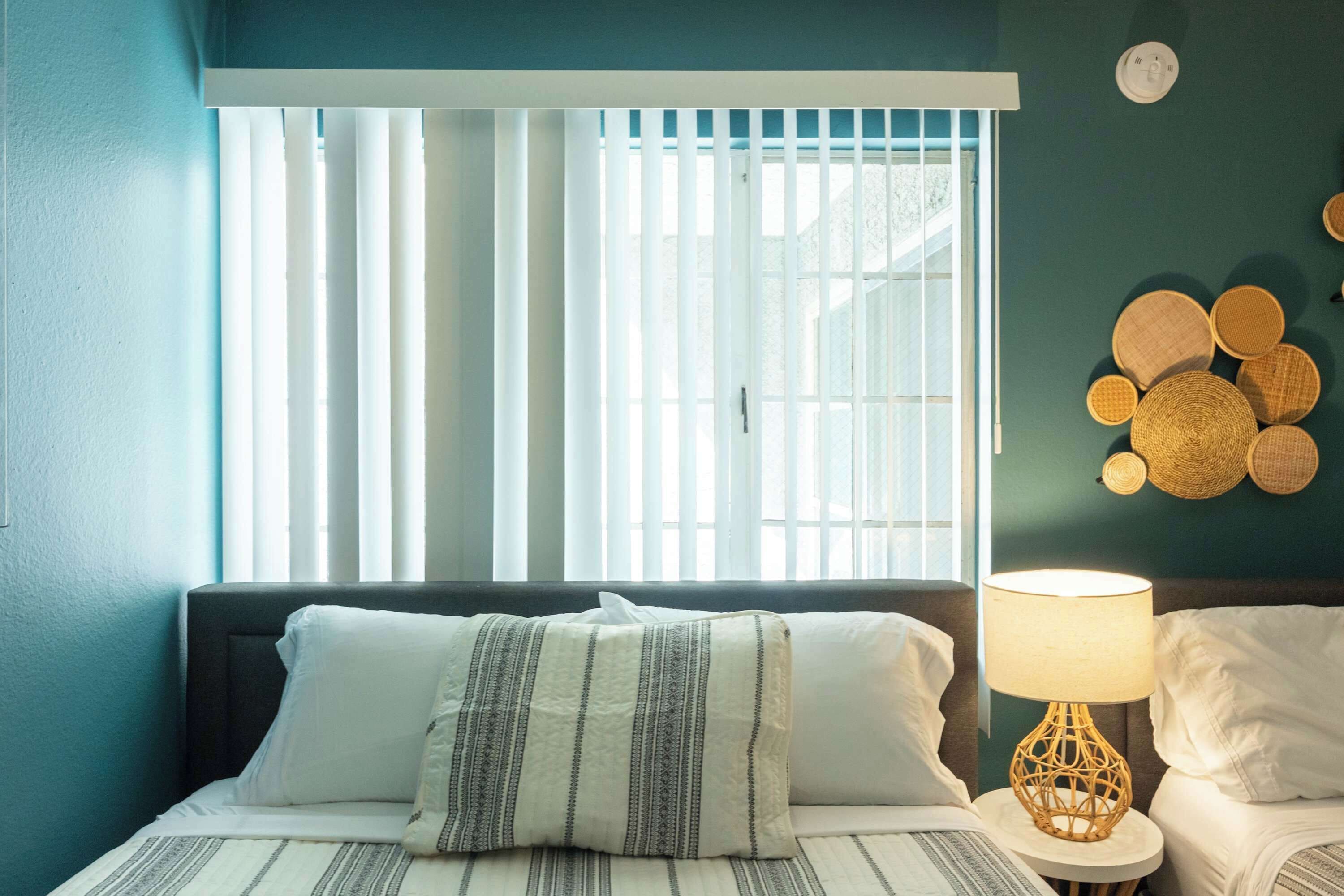 A bedroom with a bed, a bedside lamp, a window with blinds, and a wall decoration.