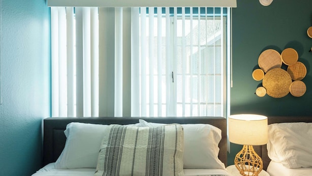 Ein Schlafzimmer mit einem Bett, einer Nachttischlampe, einem Fenster mit Jalousien und einer Wanddekoration.