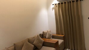 A room with a beige sofa, a wooden side table, beige curtains, and an air conditioning unit mounted on the wall. - 7 The Business Hotel (Aluva)