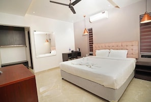 A bedroom with a bed, a desk, a chair, a ceiling fan, and a hanging light fixture. - 7 The Business Hotel (Aluva)