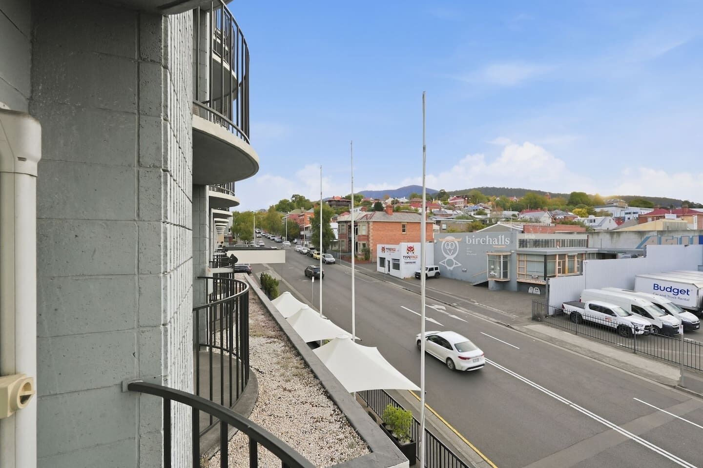 A street view with a building featuring a balcony, a road with cars, and a sign for 'birchalls.'
