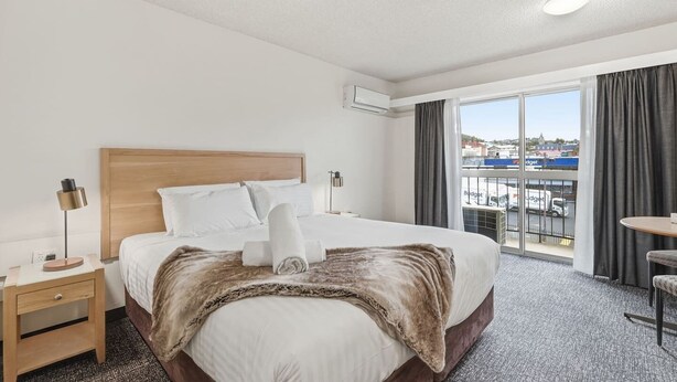 Habitación de hotel con una cama grande, una mesita de noche, una lámpara, una silla y vista a la ciudad a través de la ventana.