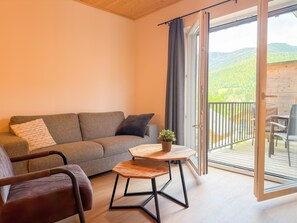 A room with a sofa, a chair, a small table with a plant, and a sliding glass door leading to an outdoor area with a table and chairs. - Cosy apartment near ski area Nassfeld (Rattendorf)