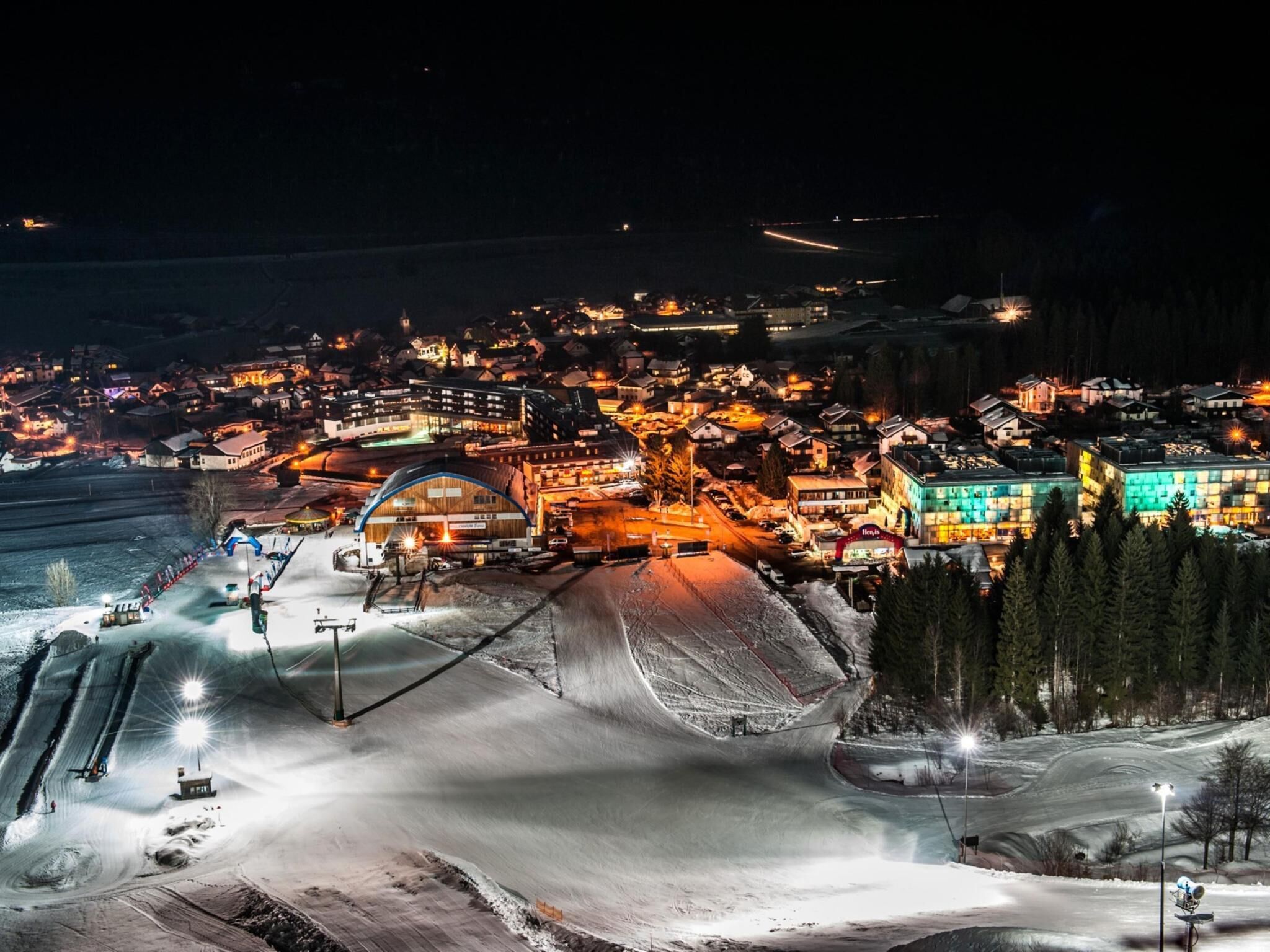 Une station de ski enneigée, avec des bâtiments illuminés, un télésiège et un remonte-pente.