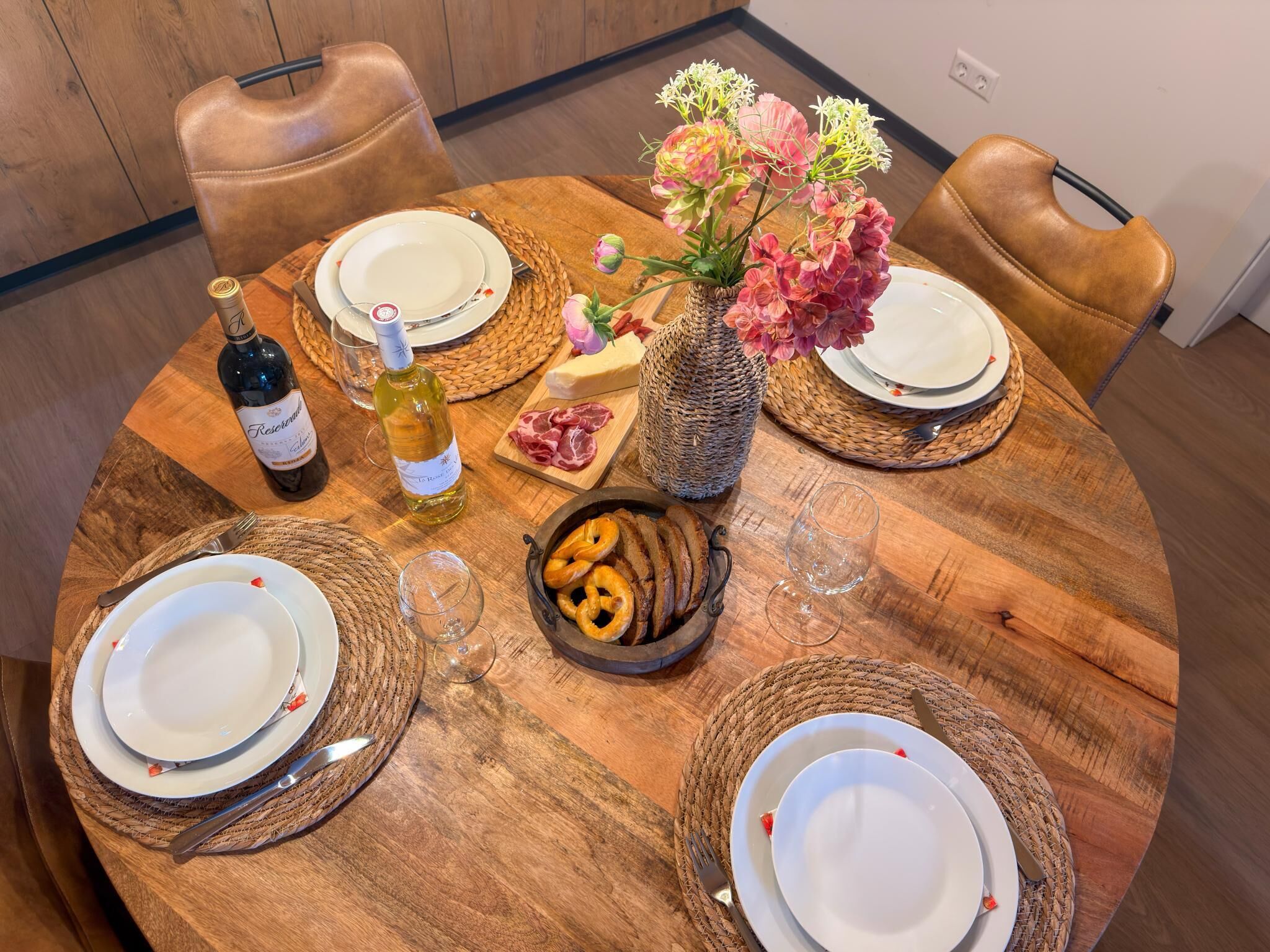 A round wooden dining table with woven placemats, plates, cutlery, a bottle of wine, a vase with flowers, and a bowl of pretzels.