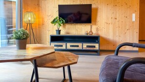A modern living room with a wooden wall, a TV mounted above a dark-colored entertainment unit, a table with a potted plant, and a chair. - Cosy apartment near ski area Nassfeld (Rattendorf)