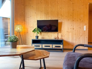 Una sala moderna con una pared de madera, un televisor montado sobre un mueble de entretenimiento oscuro, una mesa con una planta en maceta y una silla.