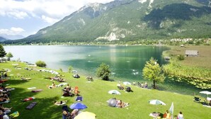 A lakeside view with mountains in the background, a grassy area with people and umbrellas, and a clear sky. - Cosy apartment near ski area Nassfeld (Rattendorf)