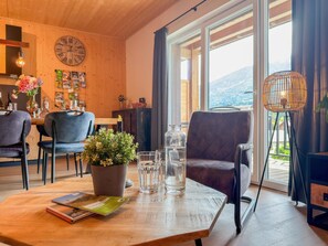 Ein gemütliches Zimmer mit Holzbalkendecke, einem Essbereich mit Stühlen und Tisch, einem großen Fenster mit Vorhängen sowie Blick ins Freie.