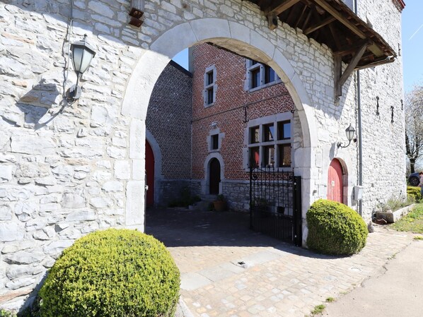 A stone archway leading to a courtyard with a brick building, a gate, and trimmed bushes.