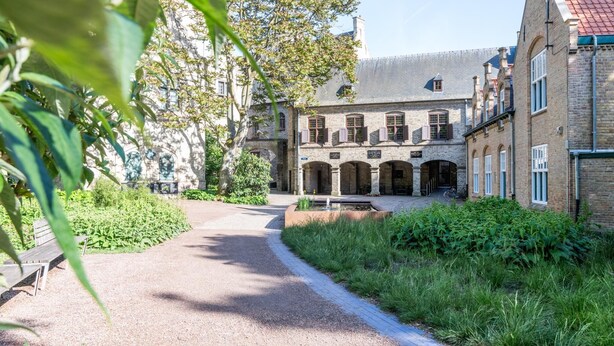 Een binnentuin met een grindpad dat leidt naar een gebouw met meerdere bogen en een rode pannendak.