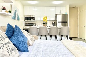 A modern bedroom with a bed, pillows, a desk, and a kitchenette. - Waikiki Sunset 1BR - FREE Parking - Walk to Beach & Zoo - AC & Pool (Honolulu)