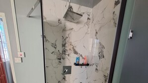 Een douchecabine met glazen wanden, een marmeren muur, een douchekop en een plankje met toiletartikelen.
