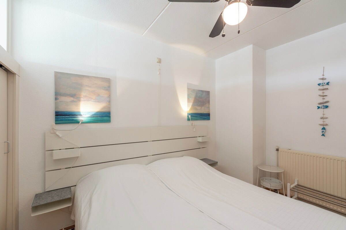A bedroom with a bed, a ceiling fan, a wall-mounted shelf, and two paintings of a sunset.