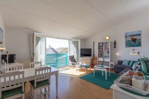 A spacious living area with a dining table, chairs, a sofa, a TV, a bookshelf, and a balcony with a view. - Bright Coastal Hideaway (Julianadorp)