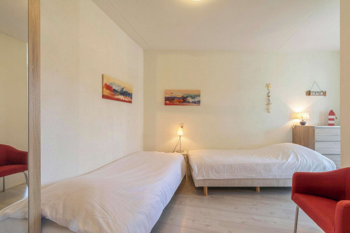 Una habitación con dos camas, una silla roja, una mesita con lámpara y decoración en la pared.