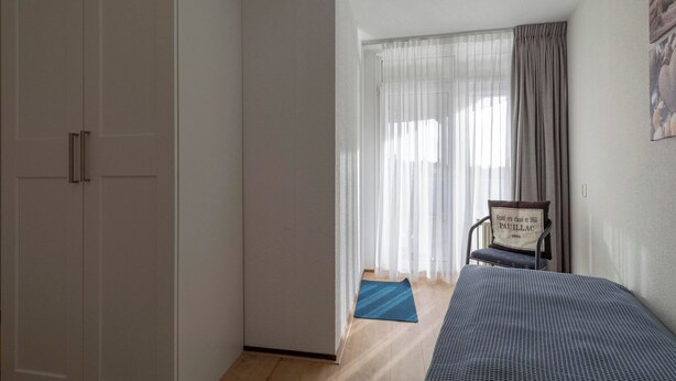 A bedroom with a bed, a chair, a wardrobe, and a window with curtains. - Cozy Dune-view Apartment (Julianadorp)