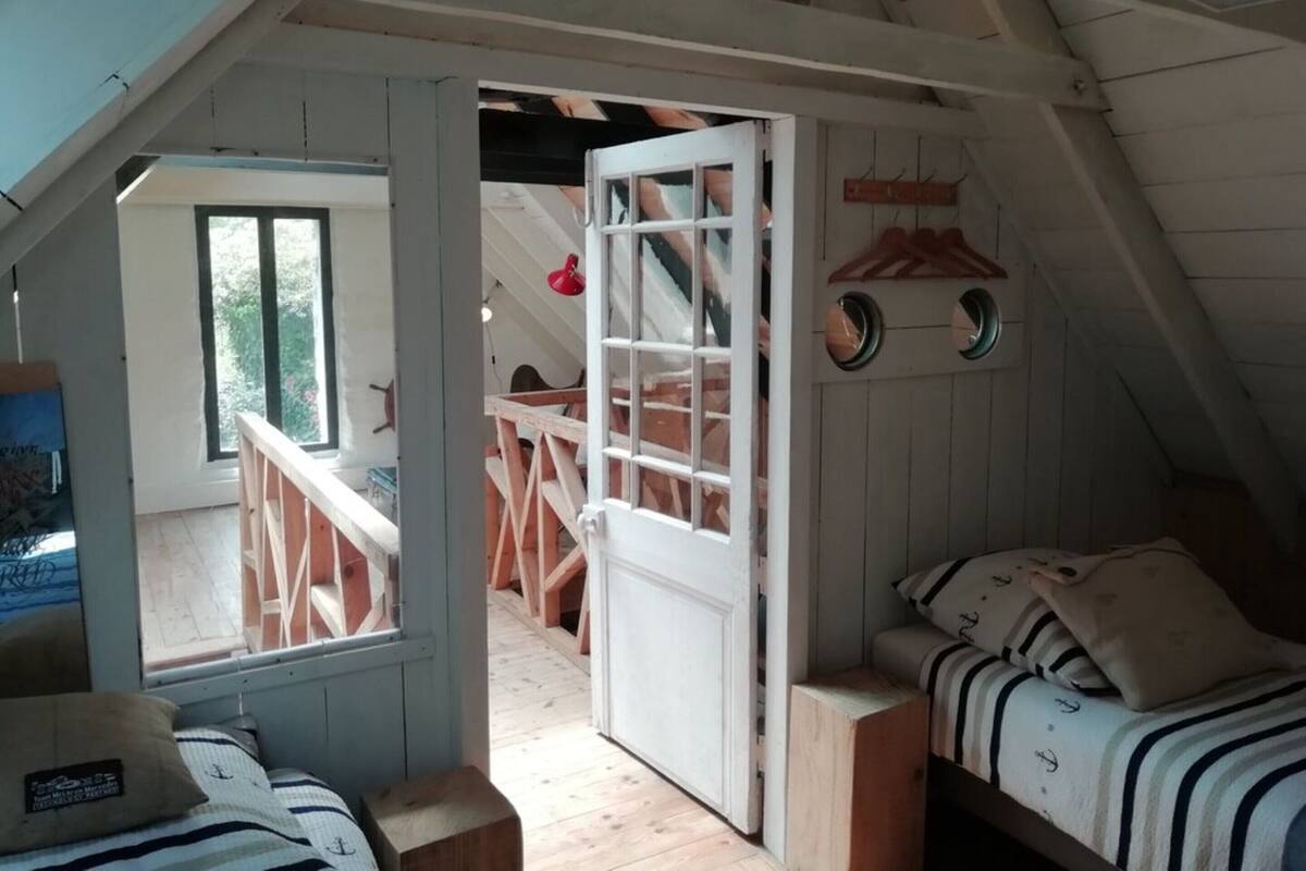 A bedroom with a wooden bed, a small bedside table, and a window. There is a door leading to another room with a wooden staircase.