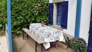 A wooden bench with a table covered by a white tablecloth with black patterns, set against a blue wall with a climbing plant and a window with blue shutters. - Brittany Escape With Garden (Cleden-Cap-Sizun)