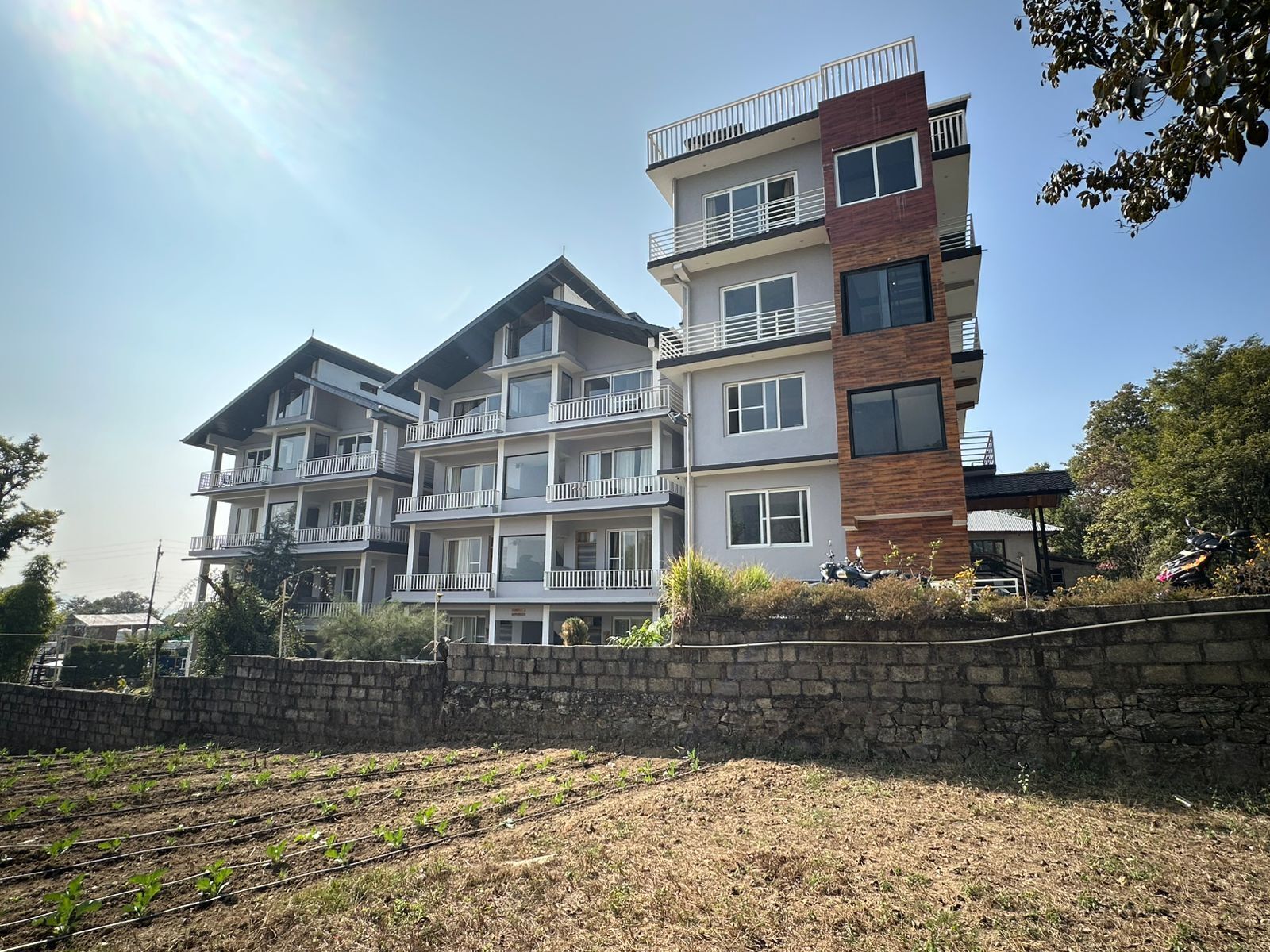 A multi-story residential building with balconies, a stone retaining wall, and a garden area with young plants.