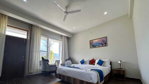 Une chambre d’hôtel avec un lit, un bureau, une chaise, un tableau, un ventilateur de plafond et une vue sur l’extérieur.