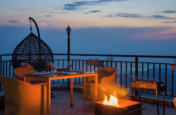 A terrace with wicker furniture, a table with wine glasses, a fire pit, and a view of the city at dusk.