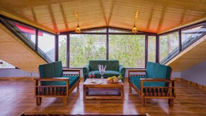 Une pièce avec un plafond en pente, un plancher en bois, un canapé vert, deux chaises en bois, une table basse avec des fruits et un vase, et de grandes fenêtres donnant sur des arbres.