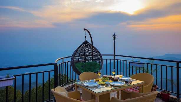 Une table à manger dressée avec des assiettes, des verres et une bouteille, entourée de chaises en osier, avec une vue sur la mer et les montagnes en arrière-plan.