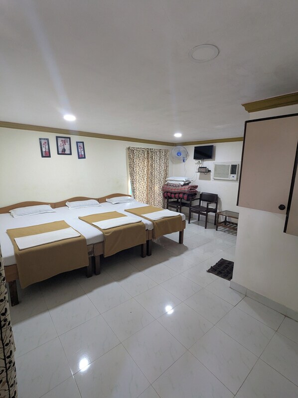 A hotel room with three beds, a desk, a chair, a TV, and a fan. - SANS RESORT (Alibag)