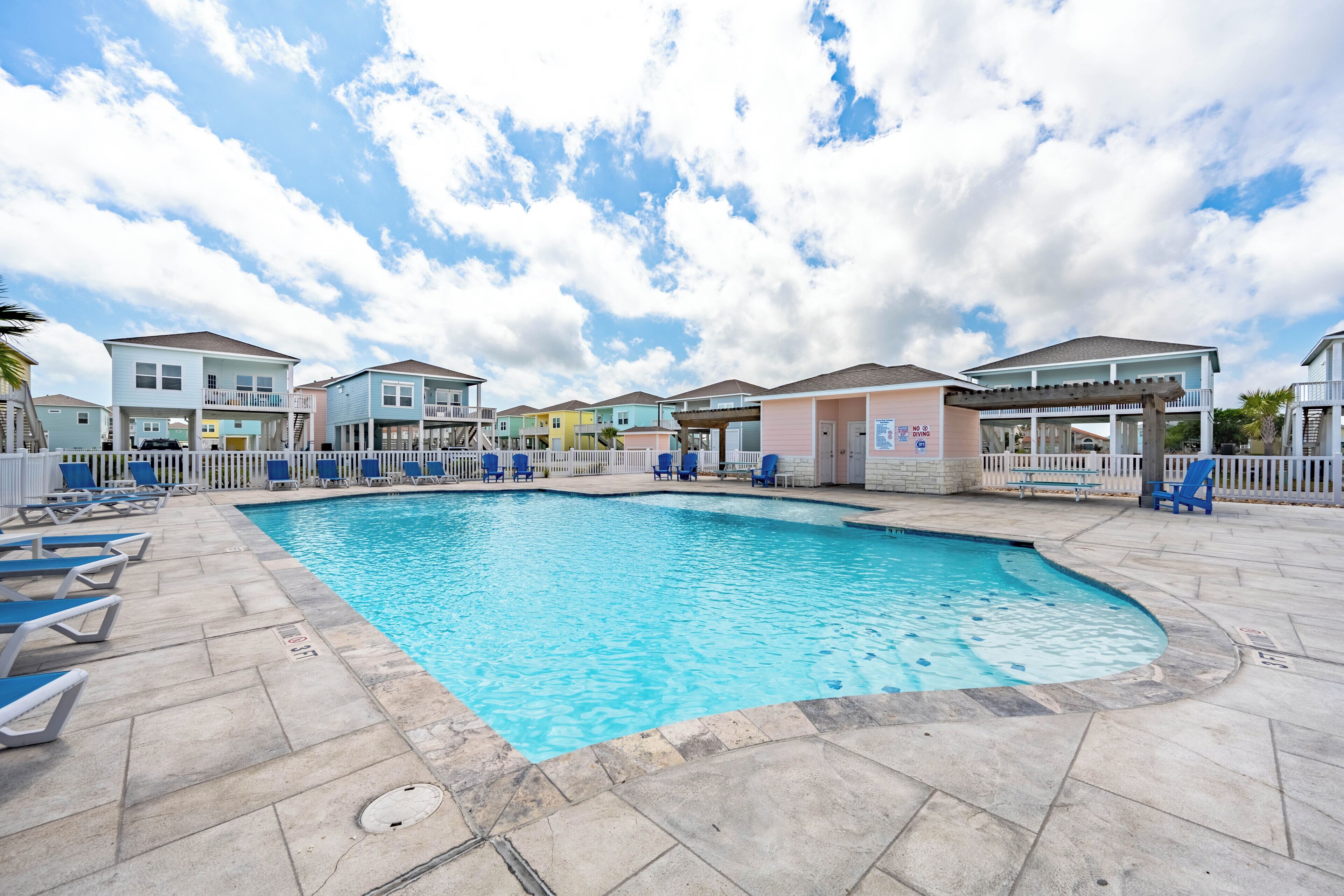A swimming pool with lounge chairs, a covered area, and colorful buildings in the background.