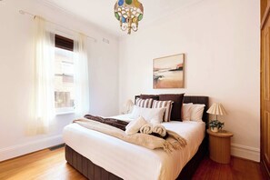 A bedroom with a bed, a bedside table with a lamp, a window with curtains, and a wall-mounted painting. - 4BR Arncliffe Hidden Gem | Bungalow with Pool+BBQ (Arncliffe)