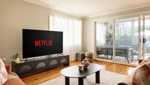 Een woonkamer met een flatscreen-tv waarop 'NETFLIX' staat, een bank, een salontafel en een schuifpui naar een terras.