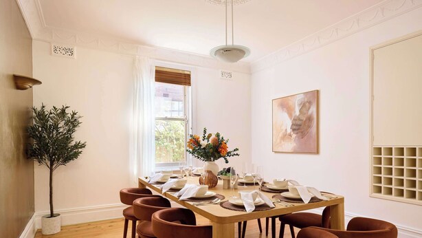 Een eetkamer met een houten tafel gedekt voor zes personen, omringd door zes bruine stoelen, een potplant en een raam met gordijnen.