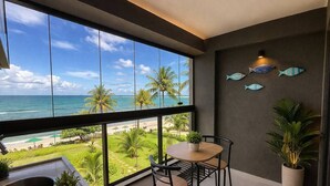 A balcony with a view of the ocean, a dining table, chairs, a potted plant, and a wall-mounted fish decoration. - Kauai Praia dos Carneiros |Apartamento Pé na Areia (Tamandaré)